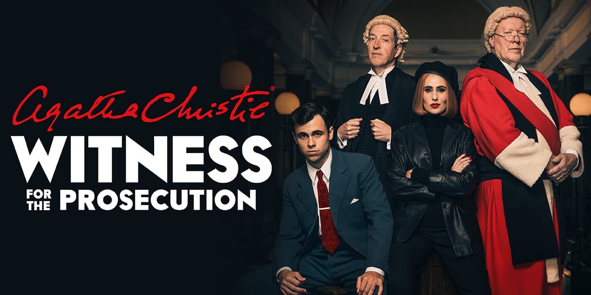 Four people stand next to text reading 'Agatha Christie Witness for the Prosecution'. Two are dressed as judges in wigs and robes, one is a woman dressed all in black, arms folded and the fourth is a man dressed in a suit. 