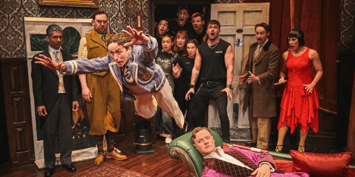 Cast of The Play That Goes Wrong on stage in a chaotic comedy scene, with one actor diving forward as others react in shock.