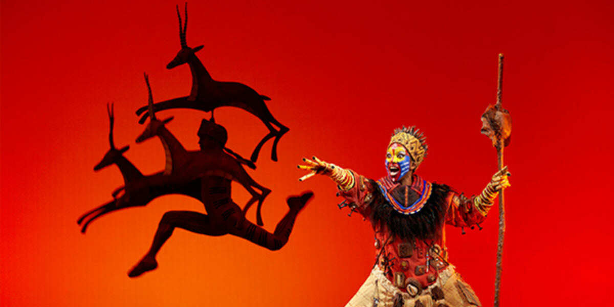 Rafiki performing on stage in The Lion King musical, with detailed costume and makeup, and shadow puppets of antelopes leaping in the background.