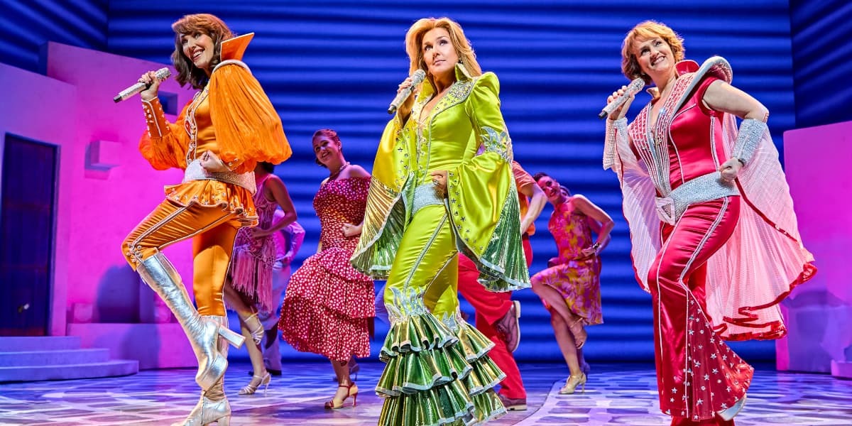Mamma Mia! musical cast performing on stage in colourful ABBA-inspired costumes, with singers in green, orange, and red flared outfits under bright stage lights.