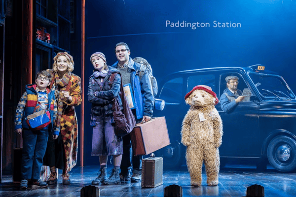 Paddington The Musical Alt Text: Image of Paddington Bear on Stage standing next to the Brown Family with a London taxi behind and a sign for Paddington Station 