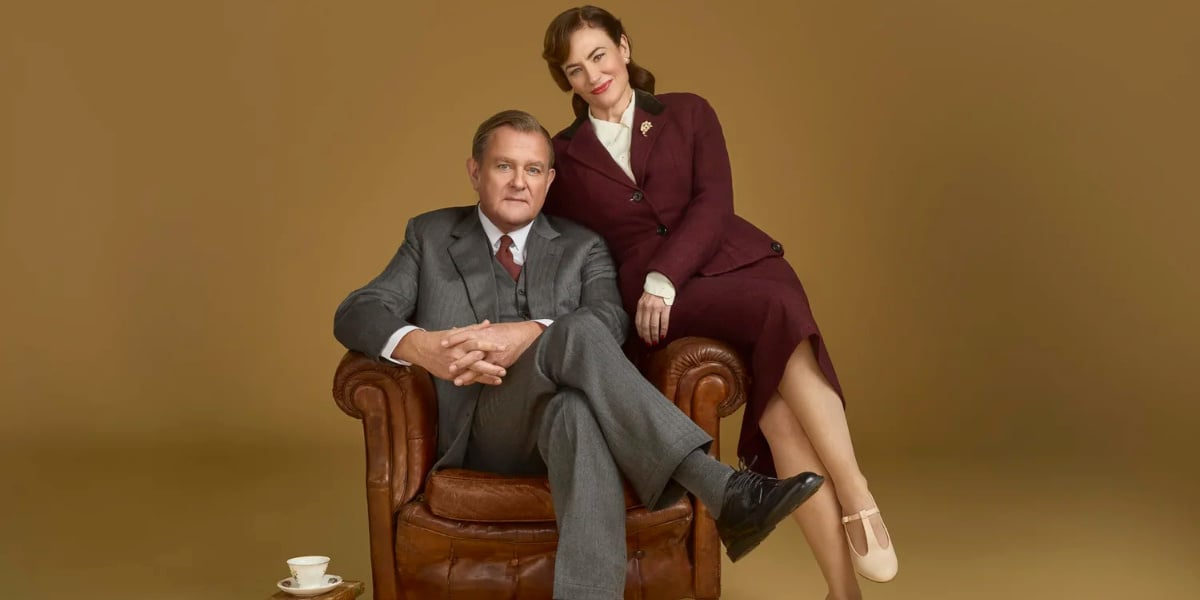 Promotional image for Shadowlands play London showing a middle-aged man seated in a leather armchair with a woman standing beside him, both dressed in 1950s-style clothing against a warm studio backdrop.