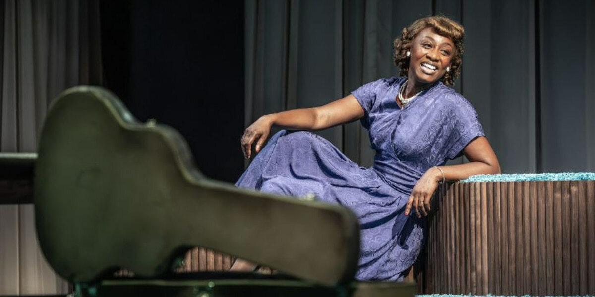 Beverley Knight on stage in a purple dress, smiling with an open guitar case in front of her in the play Marie and Rosetta, London theatre production.