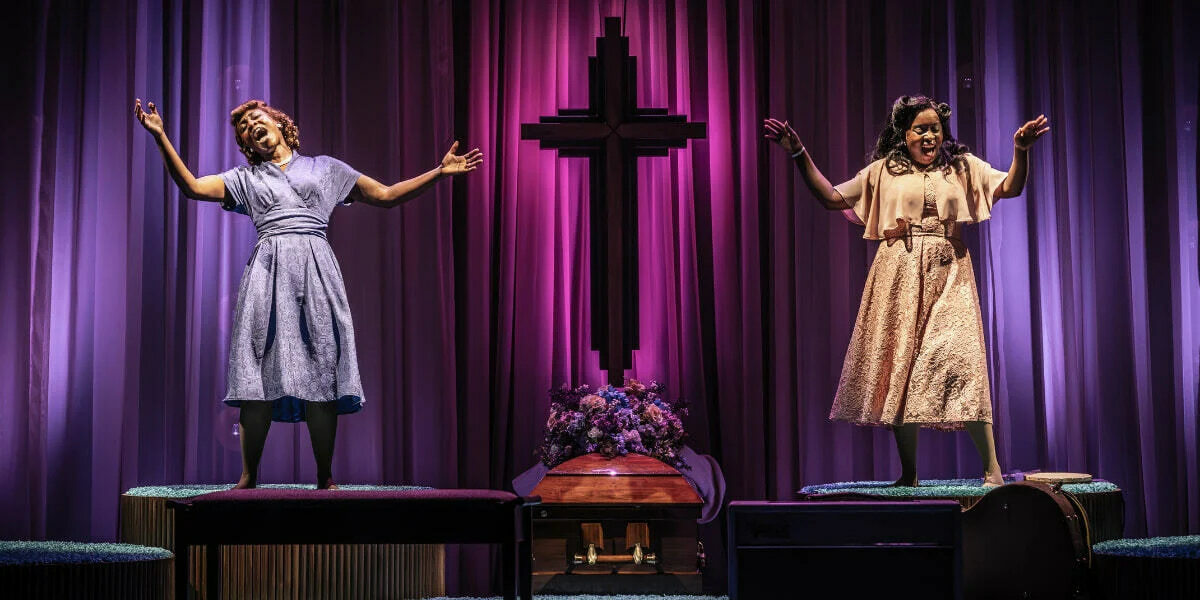 Two women sing gospel music at a funeral in Marie & Rosetta the play. The stage is lit in vibrant pink and purple hues. 