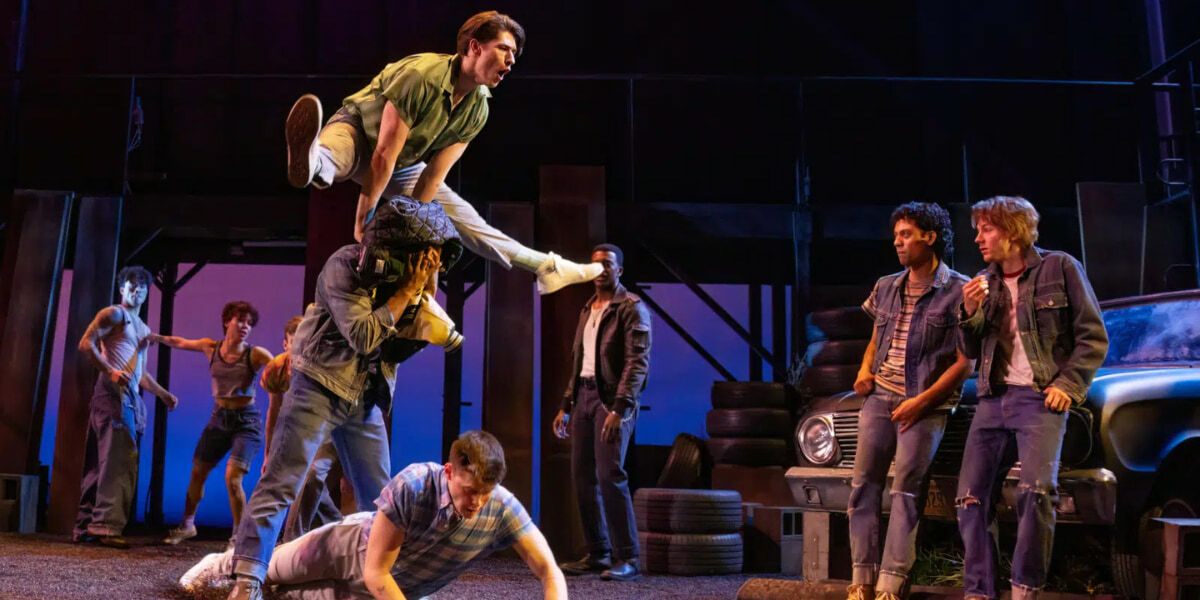 High-energy production shot from The Outsiders on Broadway showing a dramatic fight scene, with one actor leaping over another in mid-air as the ensemble reacts around a car and stacked tyres on a stylised urban stage set.