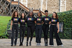 A group of six individuals stands together outdoors, all wearing matching colorful t-shirts. They are smiling and posing against a backdrop of greenery and historic architecture.