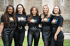 A group of five people stands together outdoors, wearing matching black T-shirts. The shirts feature colorful silhouettes of dancers.