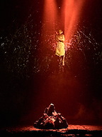 A figure in a white dress hovers above a dark, shadowy scene illuminated by red light. The atmosphere is intense, creating a dramatic visual.