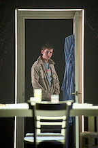 A figure stands in a doorway, looking into a dimly lit room. A table and chair are visible in the foreground.