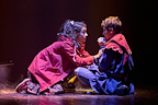 Two characters are engaged in an intense conversation on stage, surrounded by fog. They wear colorful costumes and display strong emotions.
