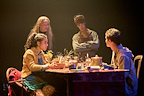 Four characters engage in conversation around a table filled with various items. The setting is dimly lit, creating a dramatic atmosphere.