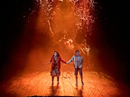 Two characters stand hand in hand on stage, surrounded by an orange spotlight. The atmosphere appears tense, suggesting a dramatic moment.