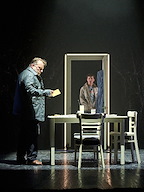 A scene from a play shows one person at a table holding a letter, while another stands in a doorway. The setting has dramatic lighting and sparse furnishings.