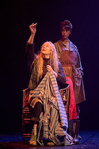 Two performers are on stage. One sits with a piece of fabric, while the other observes intently.