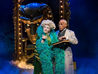 Two performers stand on stage amidst dramatic lighting. One wears a green feather boa while the other holds a book.