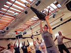 A dancer is lifted high by a partner in a rehearsal studio. Surrounding dancers cheer and celebrate.