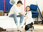 A person sits on a bathtub while another kneels beside them, looking concerned. The background features various pieces of furniture.