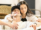 A person smiles while seated, with another person leaning affectionately on their shoulder. They are in a cozy indoor setting.