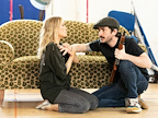 A dramatic scene unfolds between two actors on a couch. One actor kneels, holding a rifle, while the other appears distressed.
