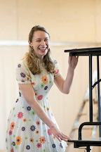 A person in a floral dress smiles while adjusting a black table. The setting appears bright and cheerful.