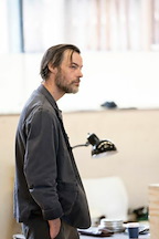 A person stands thoughtfully in an office space. They are wearing a dark jacket and are facing to the side.