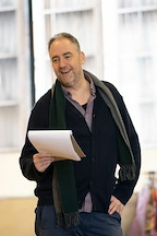 A person smiles while holding a script in a rehearsal space. They wear a dark cardigan and a scarf.