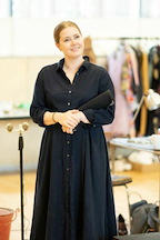 A person stands in a bright space, wearing a dark dress and holding a folder. They appear engaged and attentive.