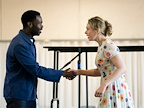 Two people shake hands while engaged in conversation, smiling warmly. The setting appears to be a rehearsal space.