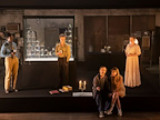 A theatrical scene features four individuals positioned around a display case filled with collectibles. They engage with one another in a dimly lit setting.