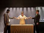 Three individuals stand around a table with candles in a dimly lit space. They are holding hands and appear to be engaged in a ritual or séance.