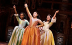 Three performers stand together in colorful, historical costumes, raising their arms in celebration. They appear to be in a theatrical setting, conveying a sense of joy and enthusiasm.