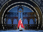 A performer in a red robe stands on stage, framed by gothic architecture. A crucifix is visible in the background.