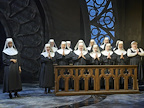 A group of individuals in religious attire performs in front of a backdrop featuring stained glass. They stand near a wooden altar with hands clasped in prayer.