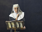 A person in a black and white habit gestures while reading from a music stand. The background is dimly lit, creating a dramatic atmosphere.