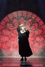 A performer in a black robe stands dramatically on stage. They hold a staff above their head against a red stained-glass backdrop.