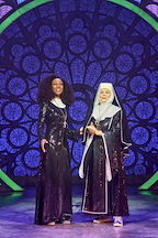 Two performers stand together on stage, smiling brightly. The background features a colorful stained glass design.