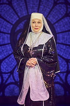 A performer stands on stage in a sparkling black and silver religious costume. The background features a stained glass design.