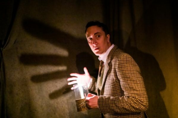 Image: Man holding a light with a shadow of his hand behind him, he is wearing a suit and has a startled/afraid look on his face. 