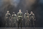 Six performers stand in military attire, posing against a smoky backdrop. The scene conveys a dramatic atmosphere.
