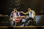 Two dancers perform on stage, one in a colorful outfit and the other in a tank top. They hold hands while creating dynamic poses.