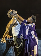 Two performers engage in a dramatic scene, one blindfolded and the other holding their face. The setting conveys a sense of tension and emotion.