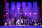 A vibrant stage performance features multiple performers in stylish attire. They are surrounded by colorful lighting and dramatic effects.