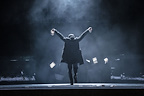 A figure stands on stage with arms raised, surrounded by swirling mist. Papers float gracefully around them.