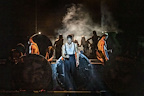 A performer stands in front of large drums, surrounded by figures in smoke. The scene conveys a dramatic atmosphere.