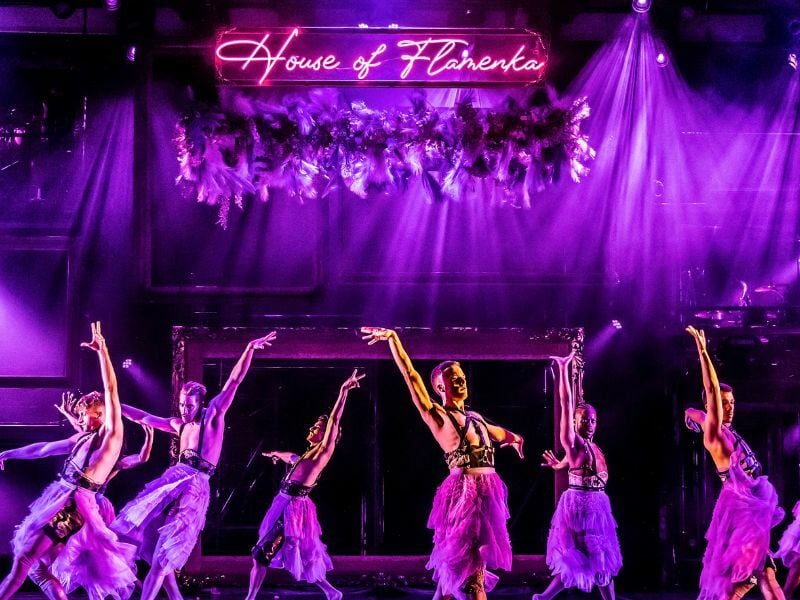 Image: cast holding their arms up, ready to dance infront of a purple lit backdrop.