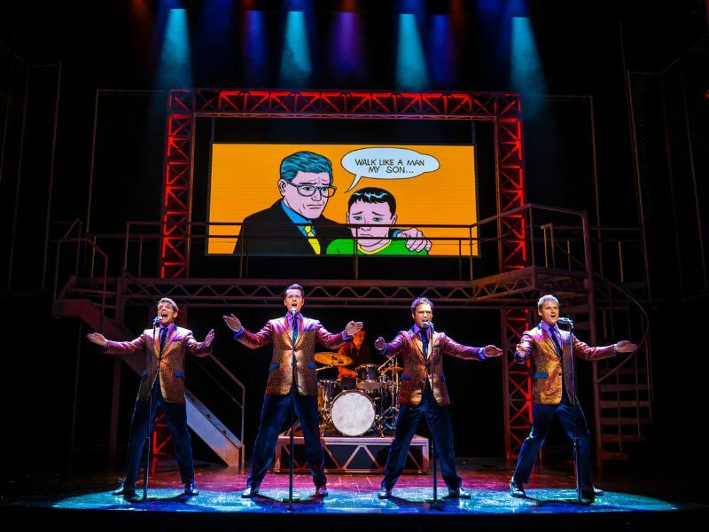 Image: the four main cast of Jersey Boys performing with their arms out. They are singing and wearing gold blazers.