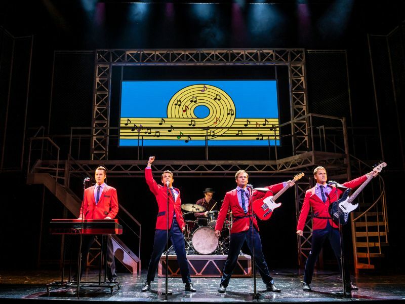 Image: the four main cast of Jersey Boys performing, wearing red blazers. They are singing and playing instruments - the guitar and the piano.