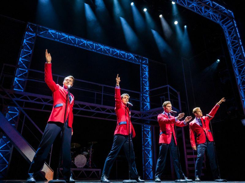Image: the cast of Jersey Boys performing on stage, the four main cast. They are wearing red blazers and are holding one arm out each.