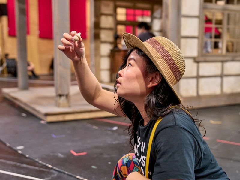Image: a female cast member wearing a hat, holding a small object up and staring at it.