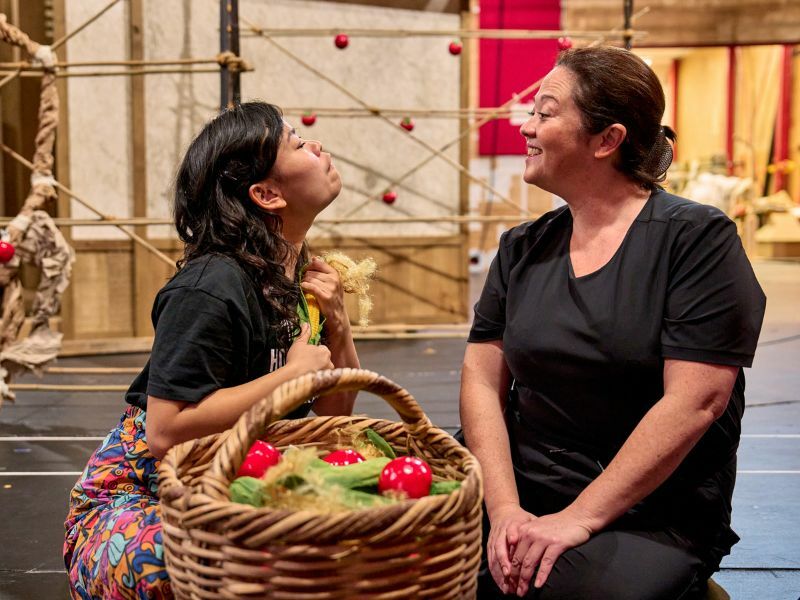 Image: two cast members interacting, one is holding a basket with fruit in.